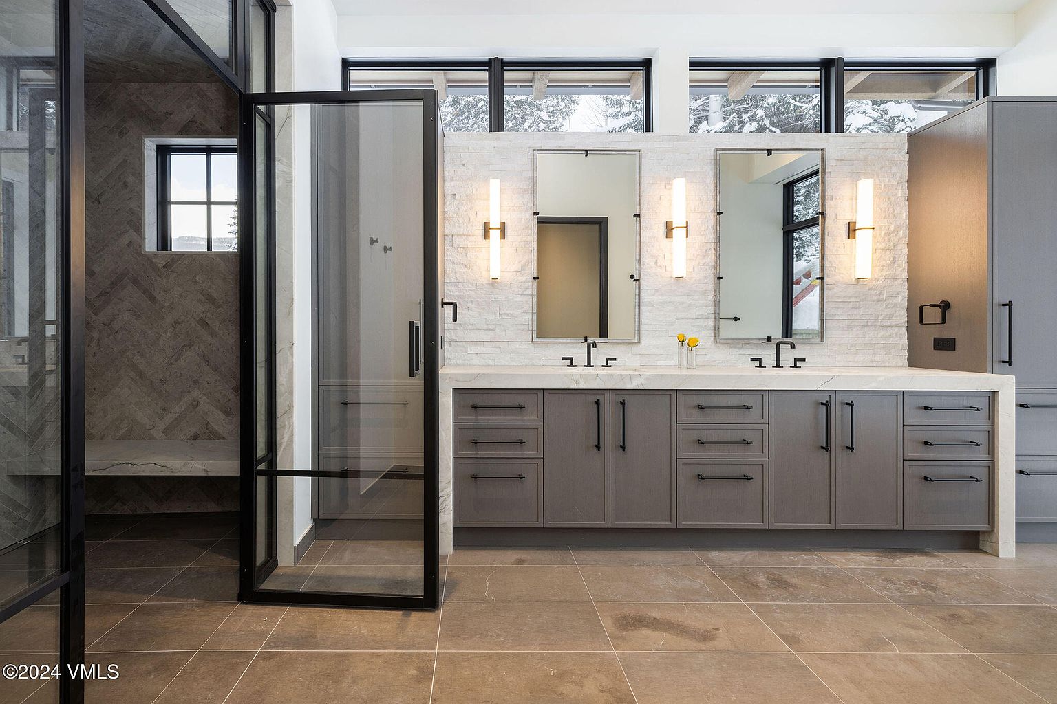 This is a primary bathroom featuring a double vanity with gray cabinetry, marble countertops, and modern black hardware. Two rectangular mirrors are mounted above the sinks, flanked by vertical sconces. A glass-enclosed shower with a bench is visible to the left, and the floor is tiled in a neutral tone. The overall impression is luxurious and contemporary.