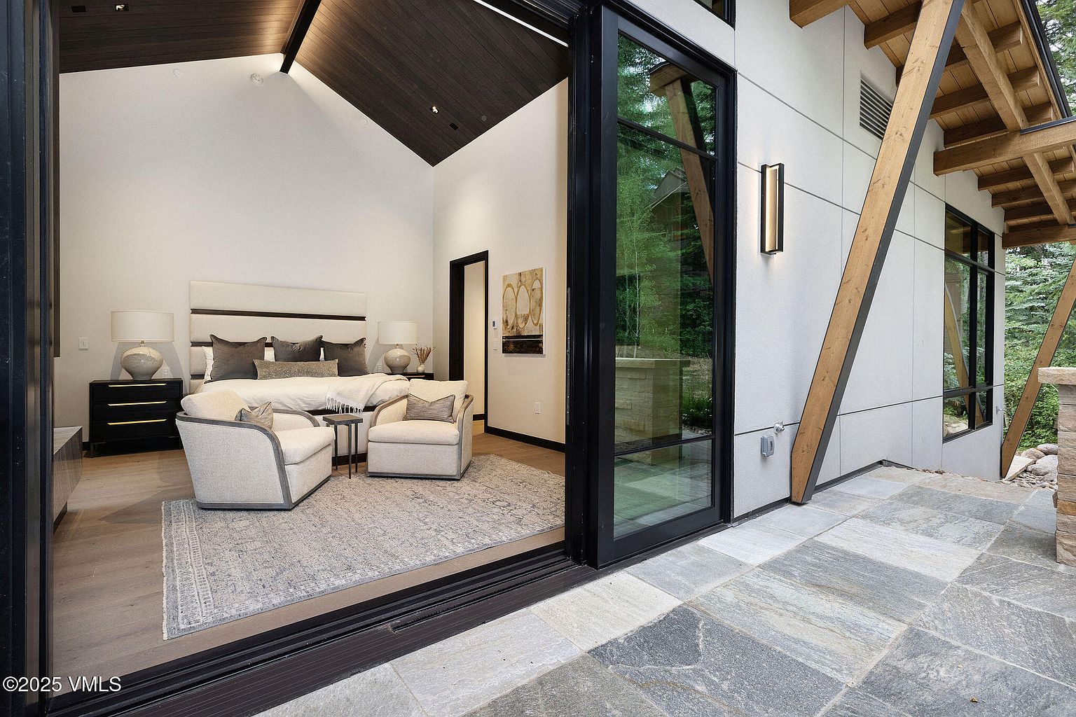 This image showcases a luxurious primary bedroom with a modern design. The room features a large bed with a padded headboard, two armchairs, and a neutral color palette. A sliding glass door opens to an outdoor patio, seamlessly blending indoor and outdoor living spaces. The high ceilings and contemporary lighting fixtures add to the room's sophisticated ambiance.