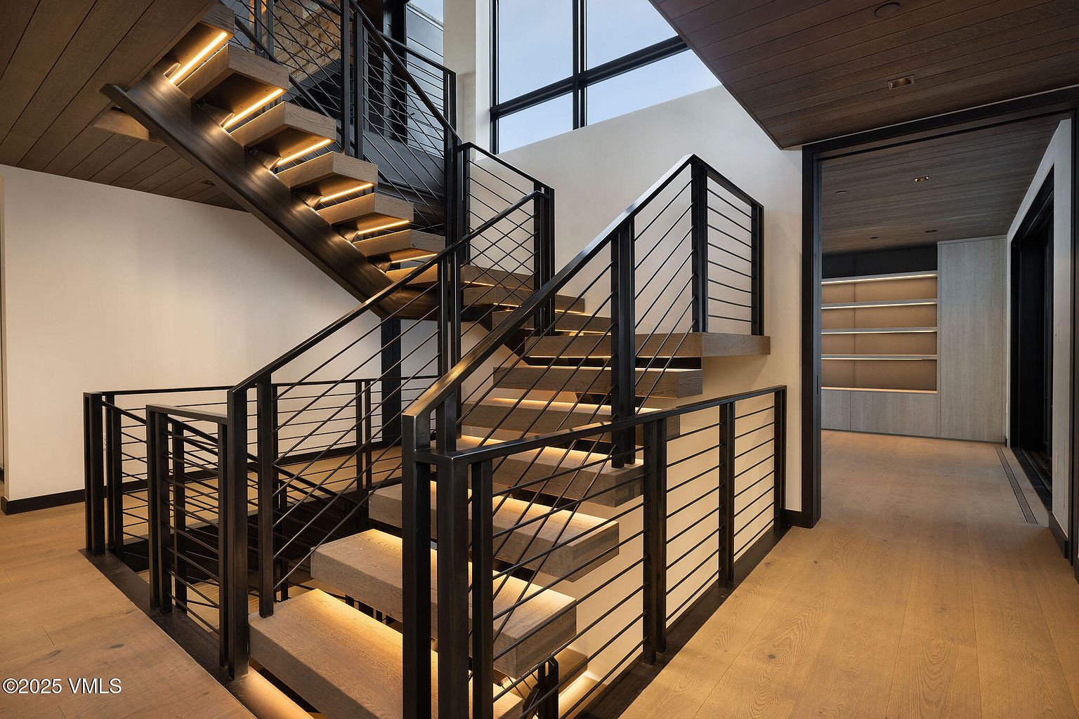 This interior shot showcases a modern staircase with wooden steps and sleek black metal railings. The steps are illuminated with subtle lighting, creating a warm and inviting ambiance. The hallway features hardwood flooring and a glimpse into another room with built-in shelving, suggesting a contemporary and well-designed home.