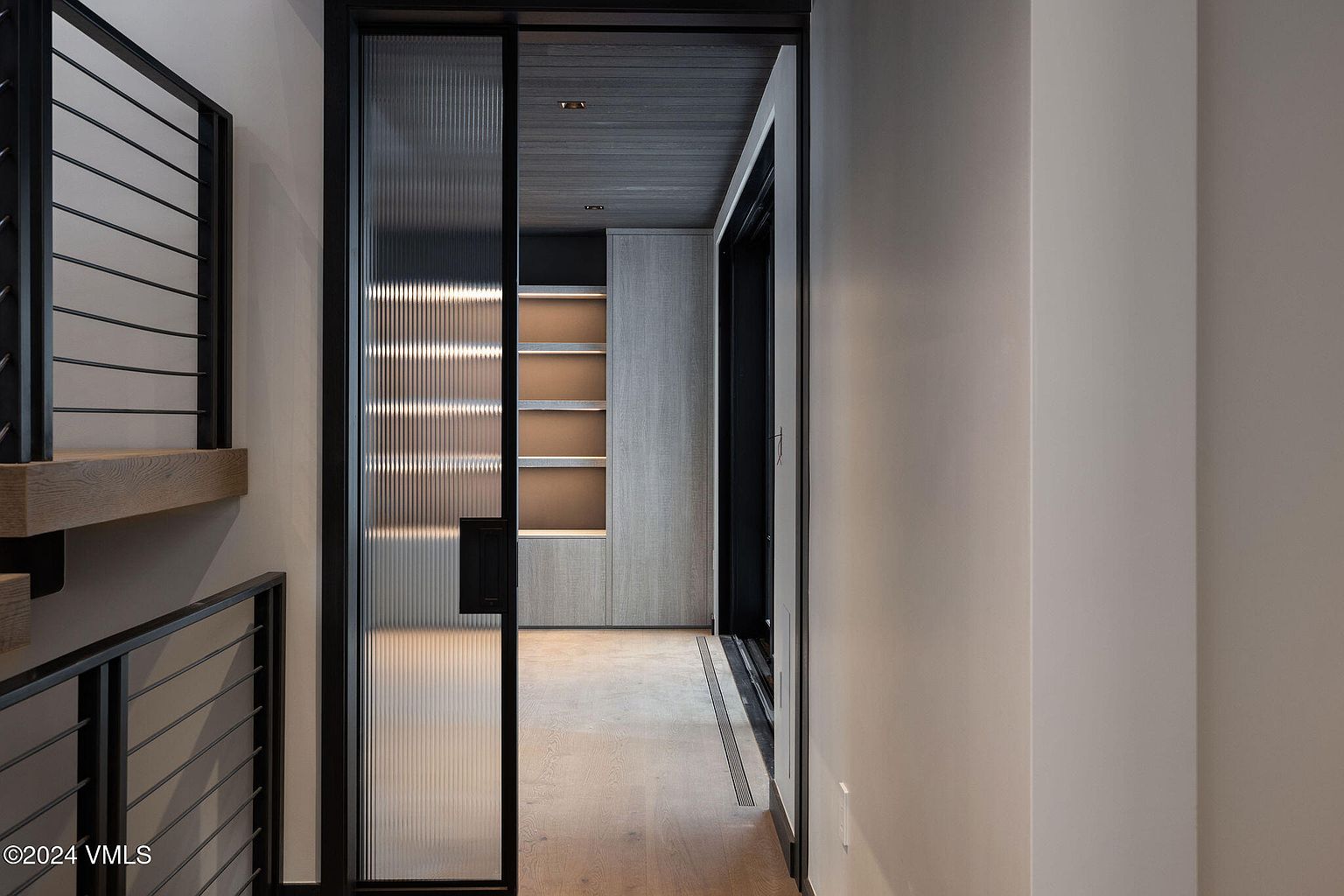 This interior shot showcases a modern hallway with a unique glass-paneled door and a glimpse into a room with built-in shelving. The hallway features light wood flooring and neutral-toned walls, creating a clean and contemporary aesthetic. A staircase with black metal railings is visible on the left, adding to the architectural interest.