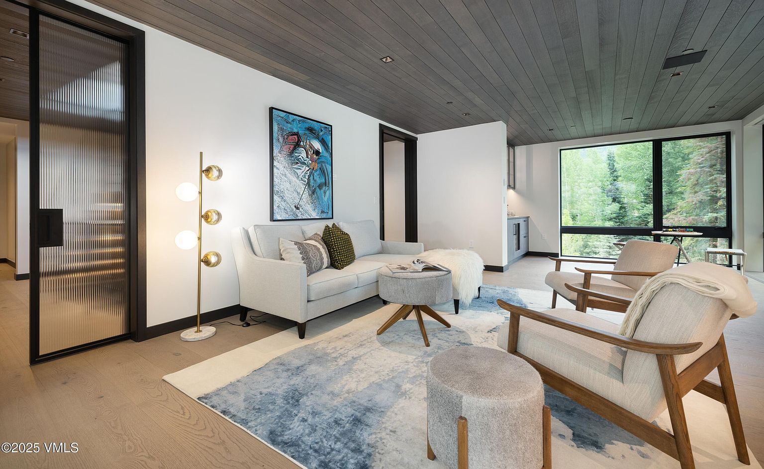 This is an interior shot of a modern living room featuring a light gray sectional sofa, two armchairs, and a blue and white area rug. The room has a contemporary design with a dark wood plank ceiling, a large window offering natural light, and a unique floor lamp. An abstract painting hangs above the sofa, adding a touch of color to the space.