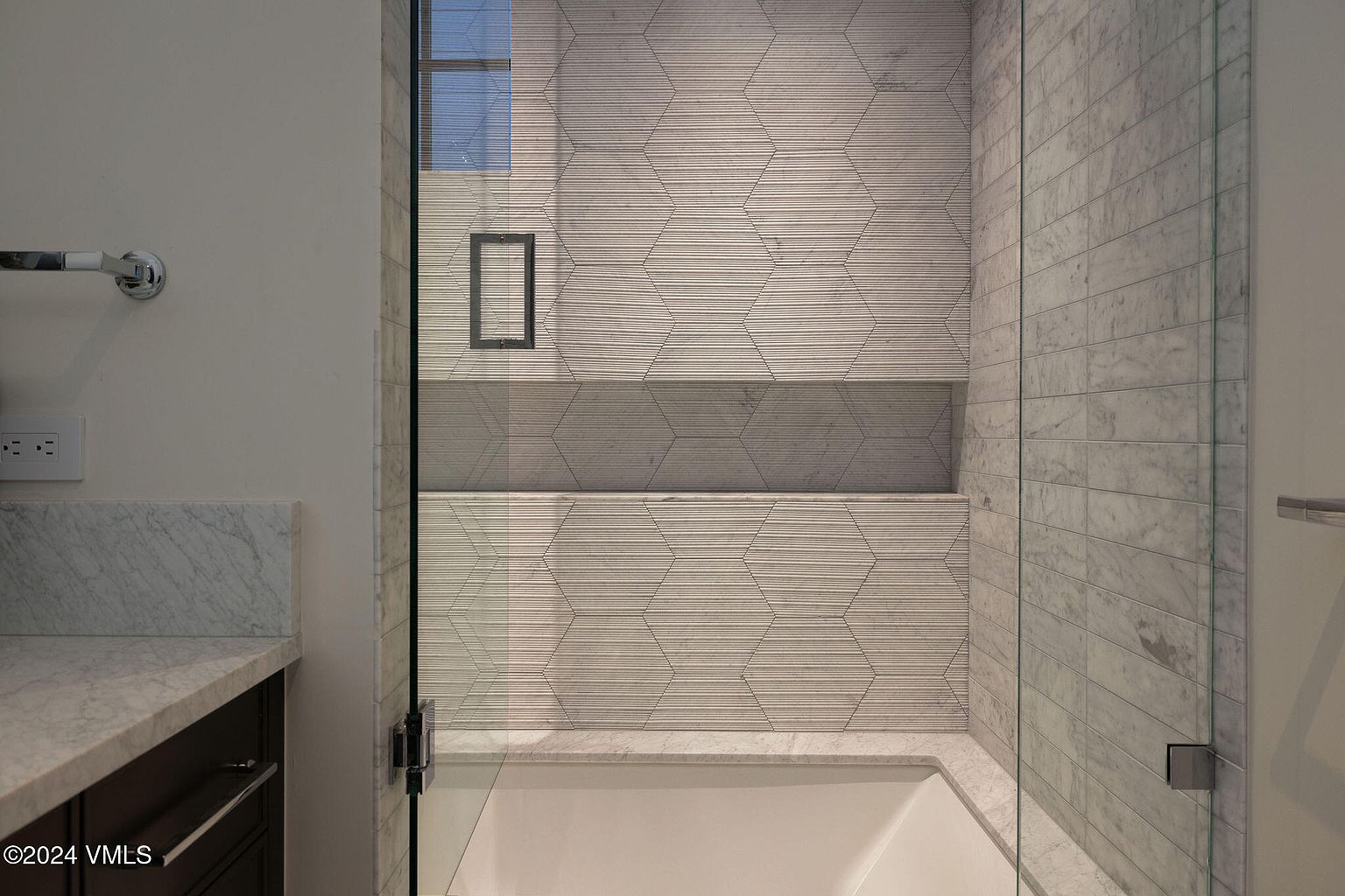 This is a detailed view of a primary bathroom shower. The shower features a glass enclosure, textured hexagonal tile walls with a built-in shelf, and marble accents. The design is modern and luxurious, emphasizing clean lines and high-end materials.