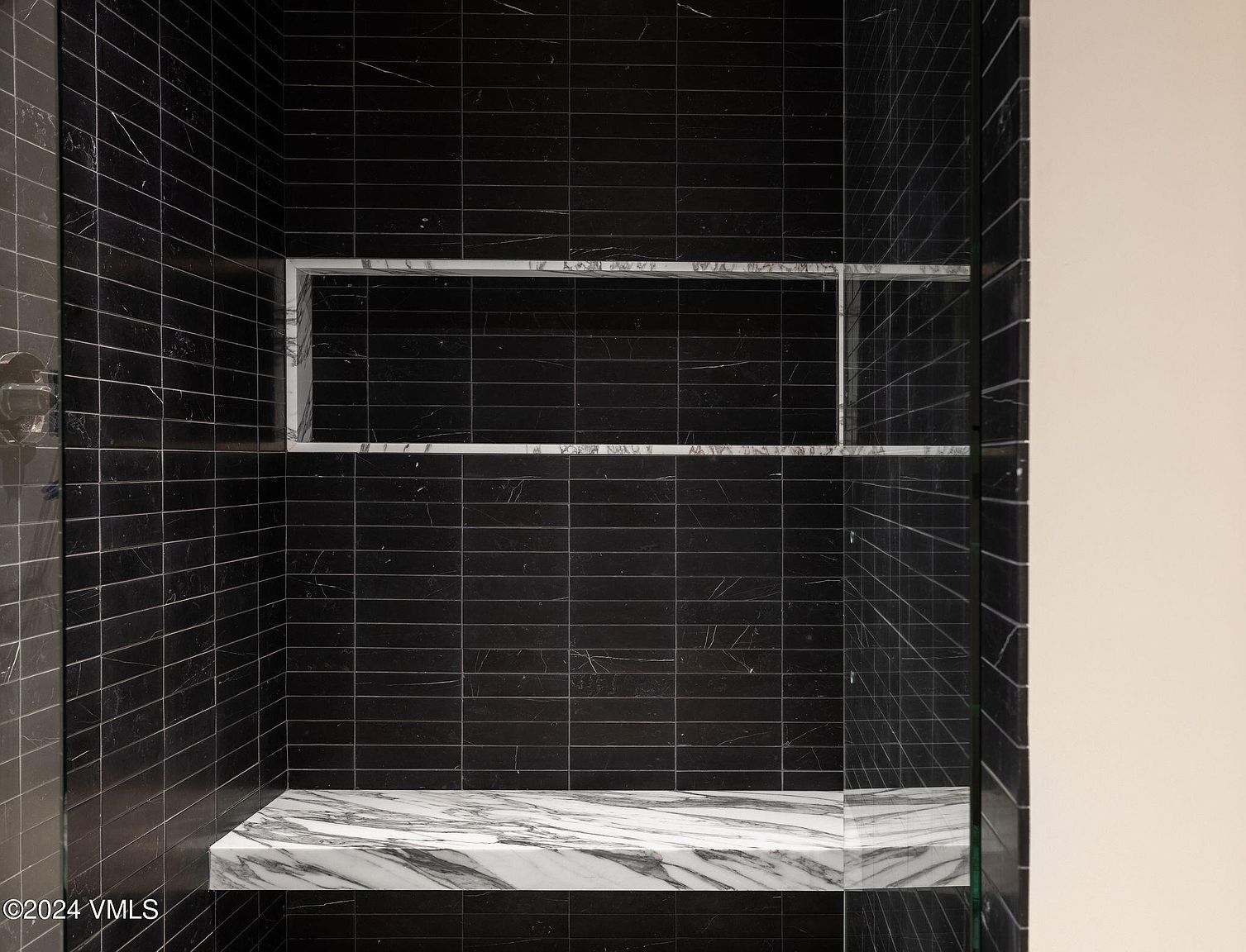 This image showcases a modern primary bathroom featuring sleek, dark tiled walls and a built-in marble bench. A recessed shelf with marble trim adds a touch of luxury and functionality. The overall design is minimalist and sophisticated, creating a spa-like atmosphere.