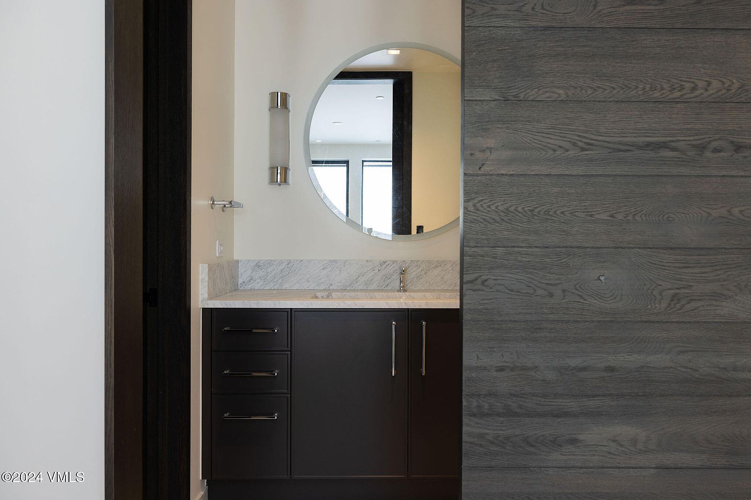 This is a modern bathroom featuring a dark wood vanity with a marble countertop and a round mirror. A sliding wood panel door adds a contemporary touch. The color palette is neutral, creating a serene and sophisticated atmosphere.