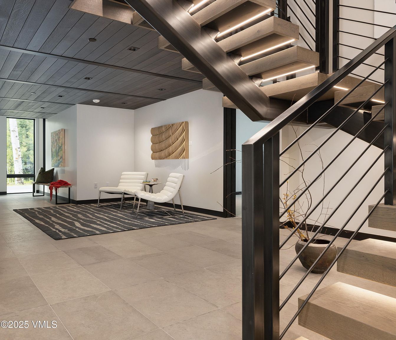 This interior shot showcases a modern hallway with a striking staircase featuring wooden steps and sleek metal railings. The hallway is decorated with contemporary art and minimalist furniture, creating a sophisticated and inviting atmosphere. The lighting is warm and accentuates the architectural details, highlighting the clean lines and open space.