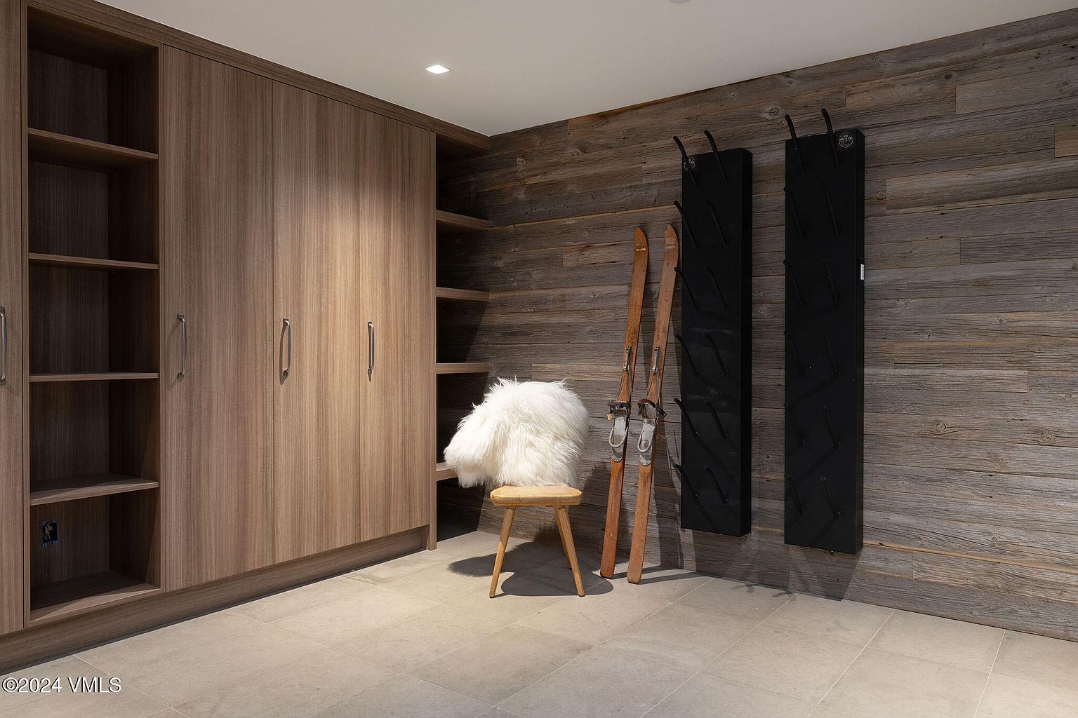 This interior shot showcases a hallway or entryway area with a rustic yet modern design. A large wooden cabinet with shelving and doors dominates the left side, while a textured wooden wall with ski racks and skis adds character to the right. A small chair with a faux fur throw sits in the middle, creating a cozy and inviting atmosphere.