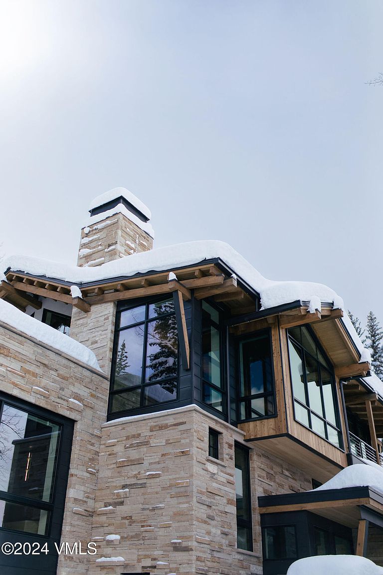 This is a low-angle, front exterior shot of a modern mountain home featuring a combination of stone and wood siding. Large windows with black frames are prominent, and snow accents the roofline and chimney, suggesting a winter setting. The architectural style is contemporary with natural elements, creating a luxurious yet rustic feel.