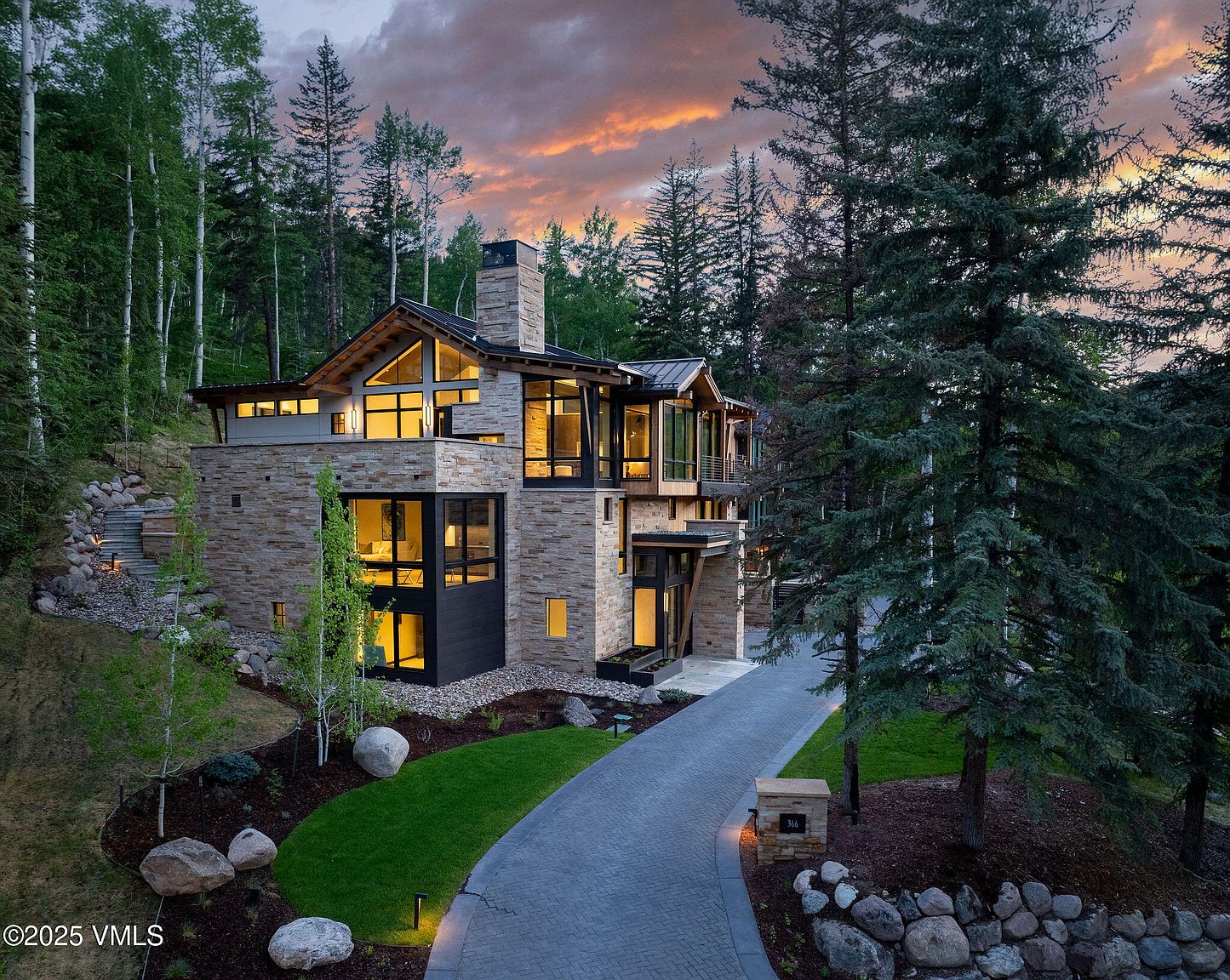 This is a stunning front view of a modern luxury home nestled in a wooded area. The exterior features a combination of stone and glass, with large windows offering ample natural light. A paved driveway leads to the entrance, and the landscaping includes mature trees, manicured lawns, and decorative rock features, creating an inviting and upscale curb appeal.