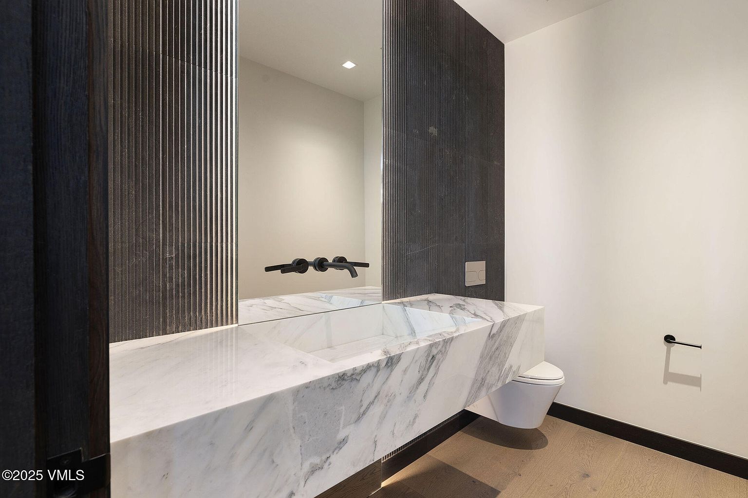 This is a modern bathroom featuring a floating marble countertop with an integrated sink and matte black fixtures. A large mirror reflects the space, and a wall-mounted toilet adds to the minimalist aesthetic. The color palette is neutral, with dark wood accents providing contrast.