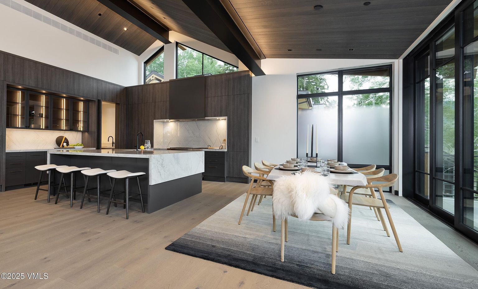 This is a modern kitchen and dining area featuring dark wood cabinetry, a marble island with seating, and a dining table set for a meal. Large windows provide natural light, and the open layout creates a spacious feel. The overall aesthetic is sophisticated and contemporary, perfect for entertaining.