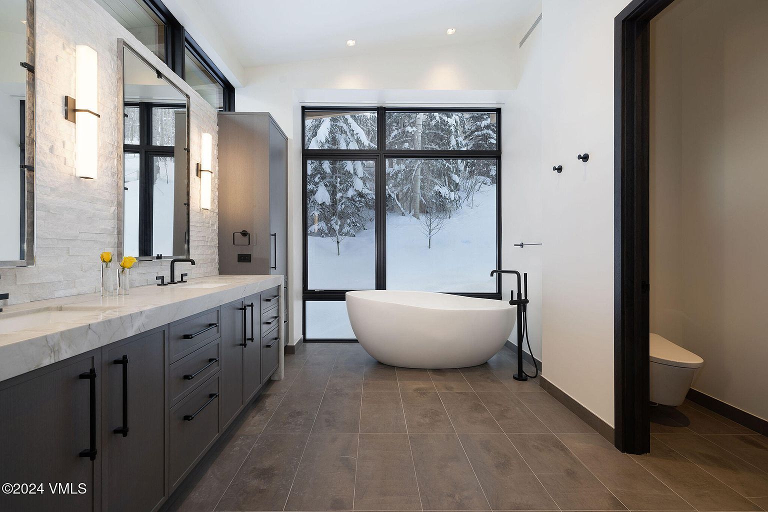 This is a primary bathroom featuring a double vanity with gray cabinets and a white countertop, complemented by modern lighting fixtures and mirrors. A freestanding white bathtub sits beneath a large window offering a view of a snowy landscape. The bathroom has a clean, contemporary design with gray tile flooring and a separate toilet area.
