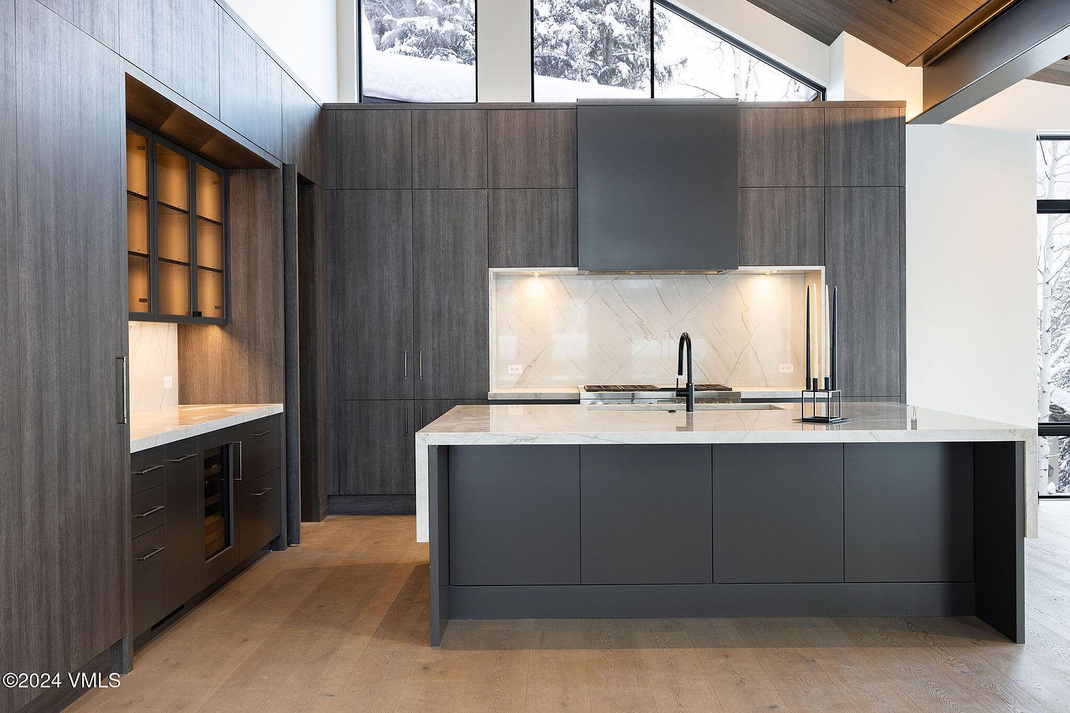 This is a modern kitchen with dark wood cabinetry and a large island with a white countertop. The kitchen features stainless steel appliances and a marble backsplash. The overall style is sleek and contemporary, with a focus on clean lines and natural materials.