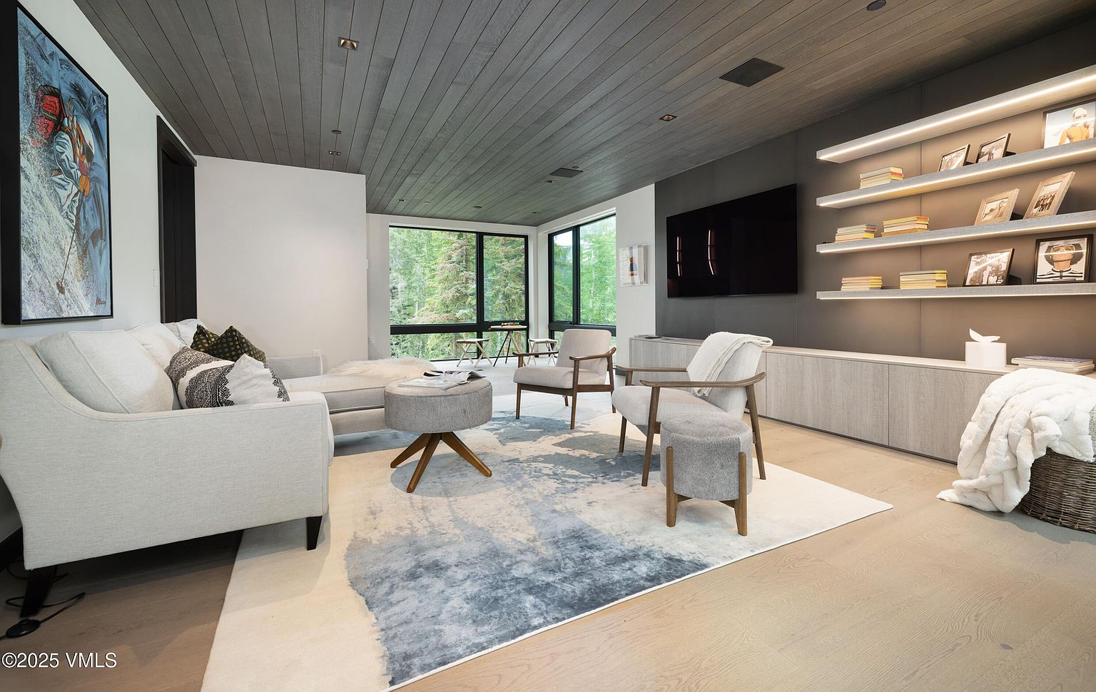 This is a modern living room featuring a light gray sofa with decorative pillows, two armchairs, and a round ottoman on a patterned rug. A large flat-screen TV is mounted on a dark gray wall with built-in shelving and accent lighting. Large windows offer a view of the surrounding greenery, and the overall style is contemporary and inviting.
