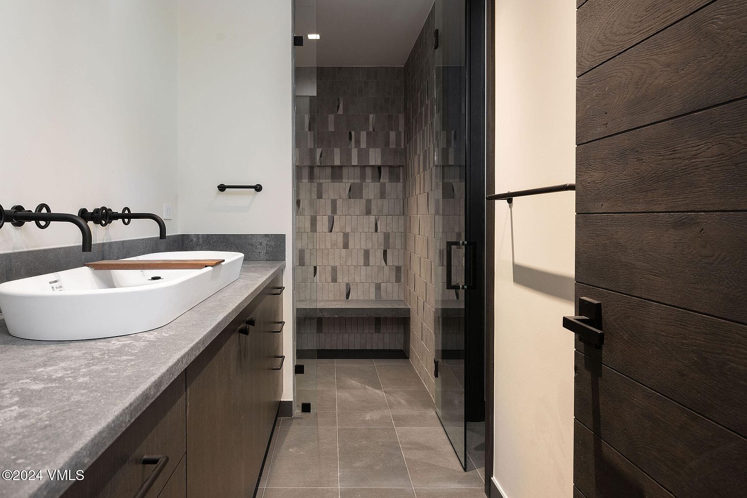 This is a modern primary bathroom featuring a double vanity with vessel sinks and matte black fixtures. The shower has unique gray tiling and a built-in bench. A dark wood door adds a touch of warmth to the otherwise cool-toned space, creating a luxurious and contemporary feel.