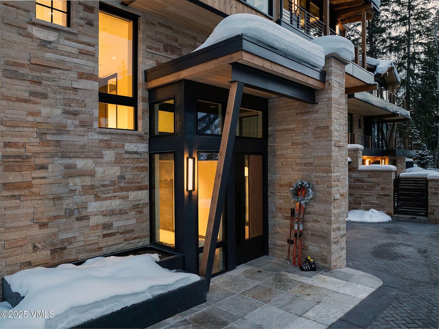 This image showcases the entryway of a modern mountain home, featuring a stone facade, a covered porch with wooden accents, and a black-framed glass door. A pair of skis and a wreath add a touch of rustic charm, while snow accents the landscape. The perspective is from a medium distance, capturing the architectural details and the welcoming ambiance of the entrance.