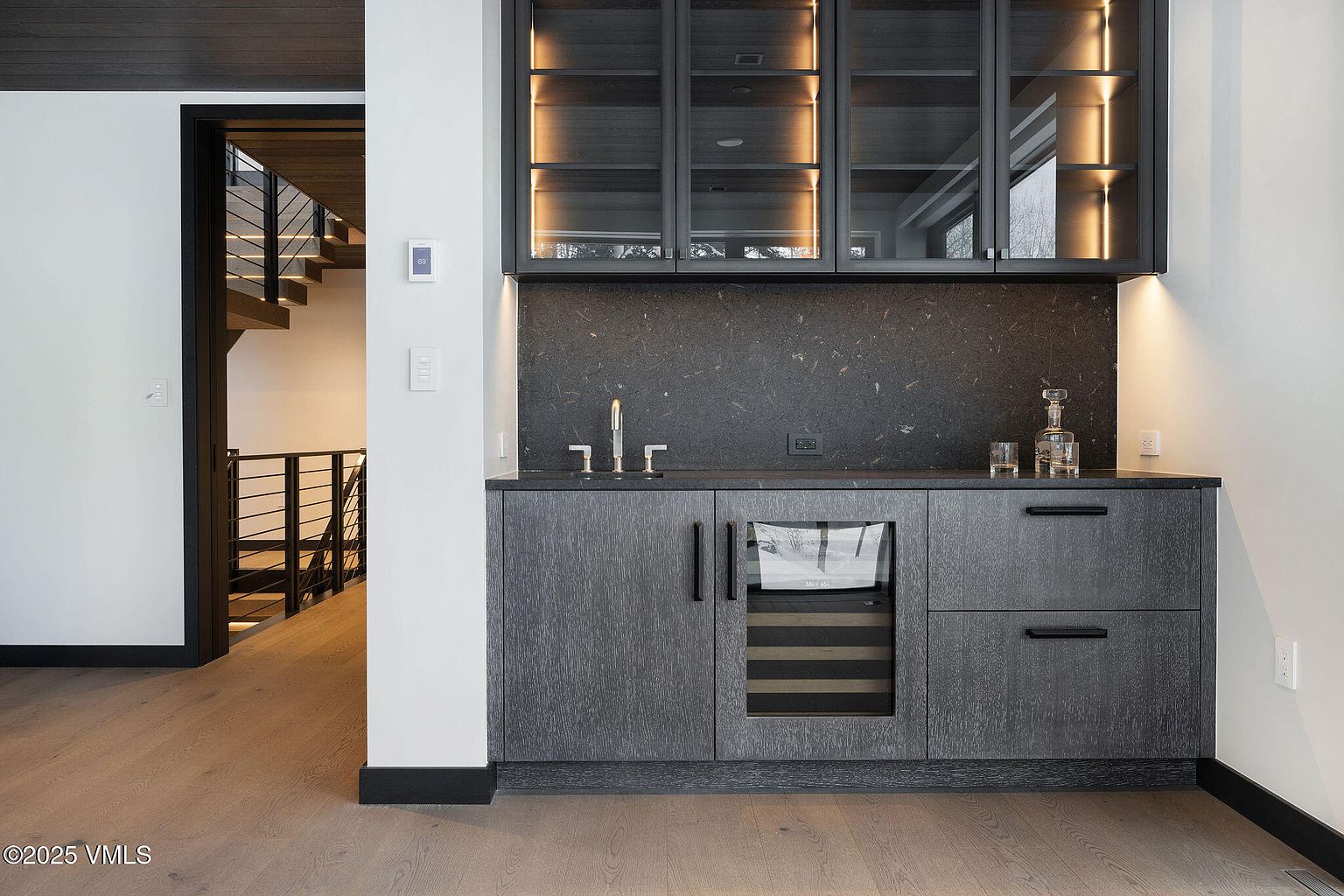 This image showcases a sophisticated wine cellar area, featuring dark wood cabinetry with glass-fronted upper cabinets illuminated by warm lighting. A built-in wine refrigerator is integrated into the lower cabinets, complemented by a dark countertop and backsplash. The overall design exudes a modern and elegant aesthetic, perfect for wine enthusiasts.