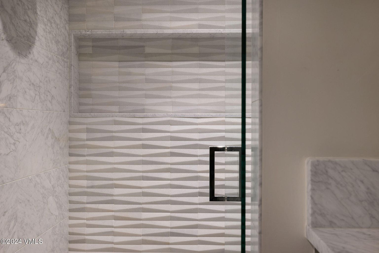 This image showcases a modern primary bathroom featuring a shower with textured, geometric-patterned tiles and a built-in niche. A glass shower door with a sleek, square handle is partially visible, adding to the contemporary aesthetic. The overall impression is clean, stylish, and luxurious.