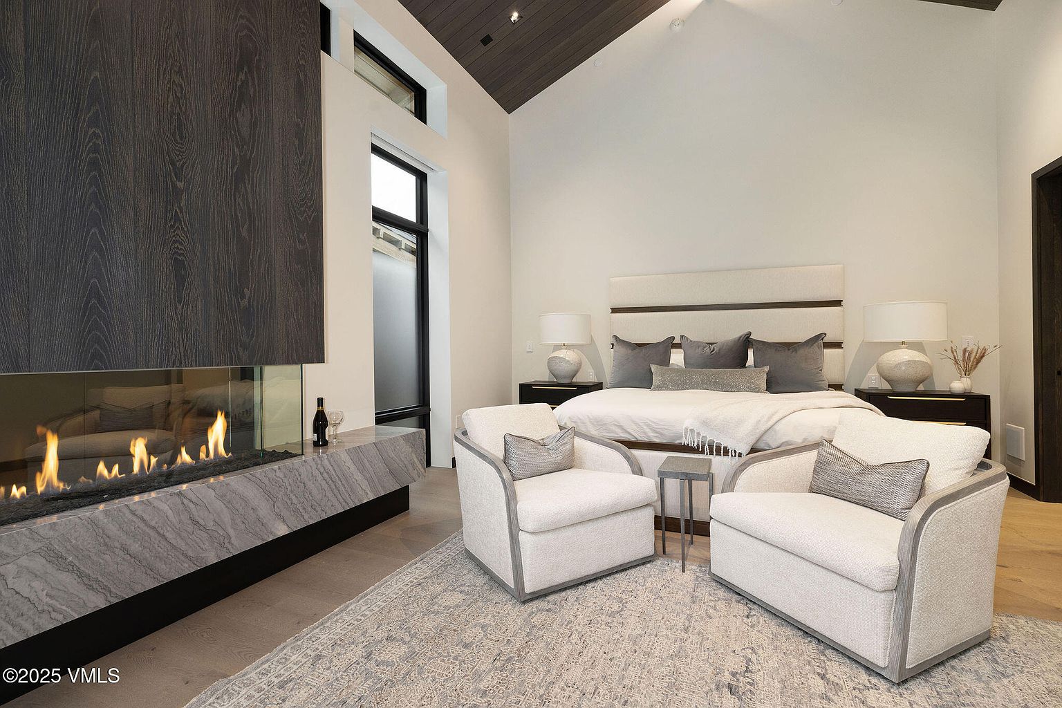 This is a primary bedroom featuring a modern fireplace with a dark wood and marble surround. The room has high ceilings and a neutral color palette, with two armchairs facing the bed. The overall impression is luxurious and serene, perfect for a relaxing retreat.