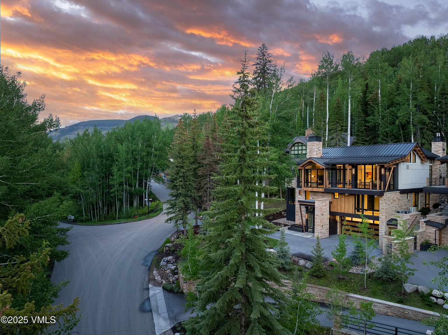 This image showcases the front exterior of a modern mountain home at sunset. The house features a combination of stone and wood siding, large windows, and a metal roof. The property is surrounded by lush trees, and a paved driveway leads to the entrance, creating a sense of privacy and luxury.