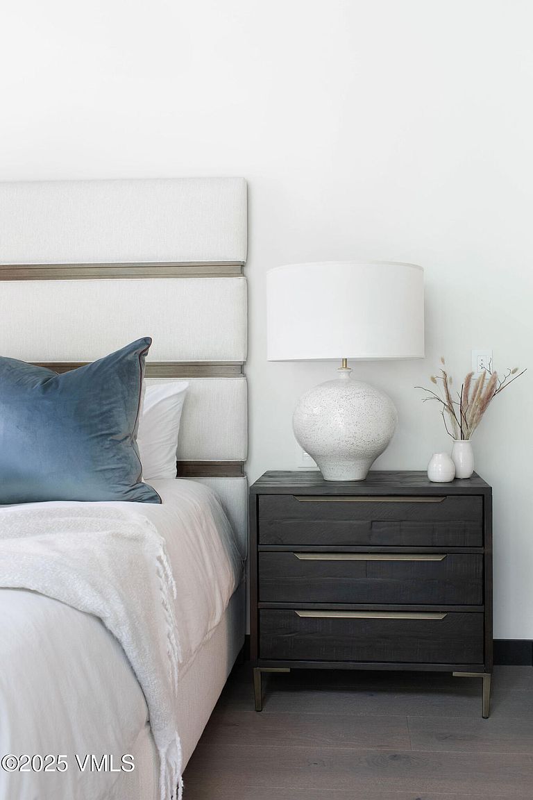 This is a bedroom interior showcasing a well-appointed bedside arrangement. A plush bed with a blue pillow and white blanket is positioned next to a dark wood nightstand, which features three drawers with sleek hardware. A white lamp and decorative vases adorn the nightstand, creating a cozy and inviting atmosphere.