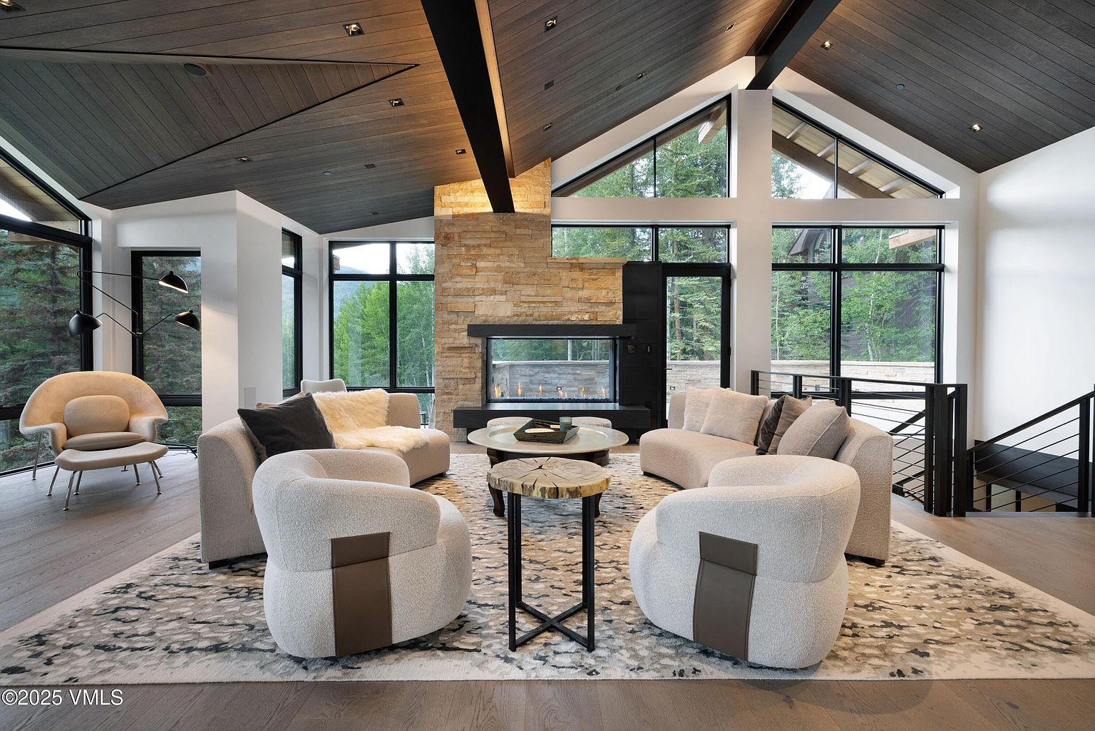 This is a spacious living room featuring a modern design with a stone fireplace and large windows offering natural light and views of the surrounding greenery. The room is furnished with curved sofas and armchairs arranged around a central coffee table, creating a cozy and inviting atmosphere. The dark wood ceiling with black beams adds a touch of sophistication and complements the neutral color palette of the furniture and rug.