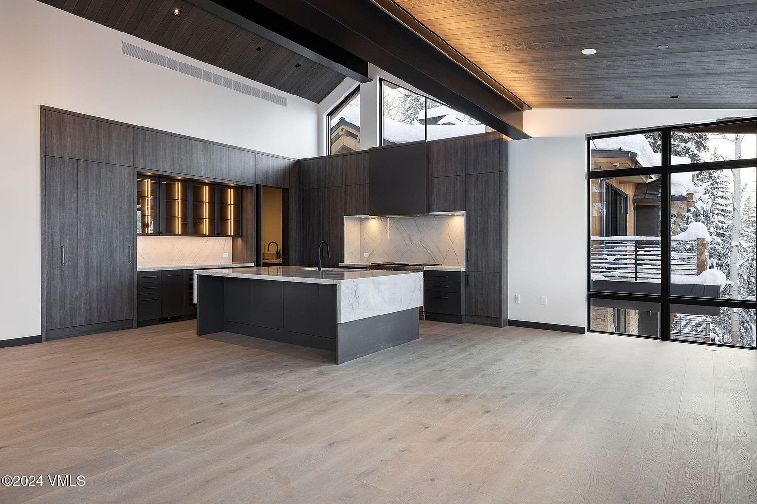 This is a modern kitchen with dark wood cabinetry and a large island with a white marble countertop. The kitchen features a sleek design with integrated appliances and a minimalist aesthetic. Large windows provide natural light and views of the snowy exterior, enhancing the contemporary and luxurious feel of the space.