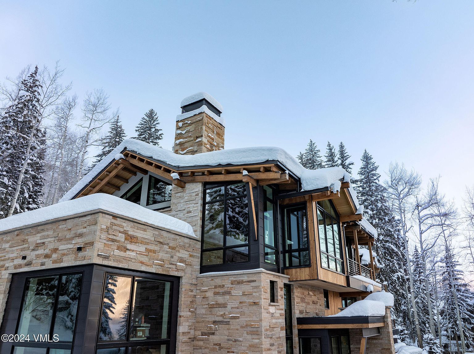 This is a front exterior view of a modern luxury home in a snowy, wooded setting. The house features a combination of stone and wood siding, large windows with black frames, and a chimney covered in snow. The architecture is contemporary with clean lines and a focus on natural materials, creating a sophisticated and inviting appearance.