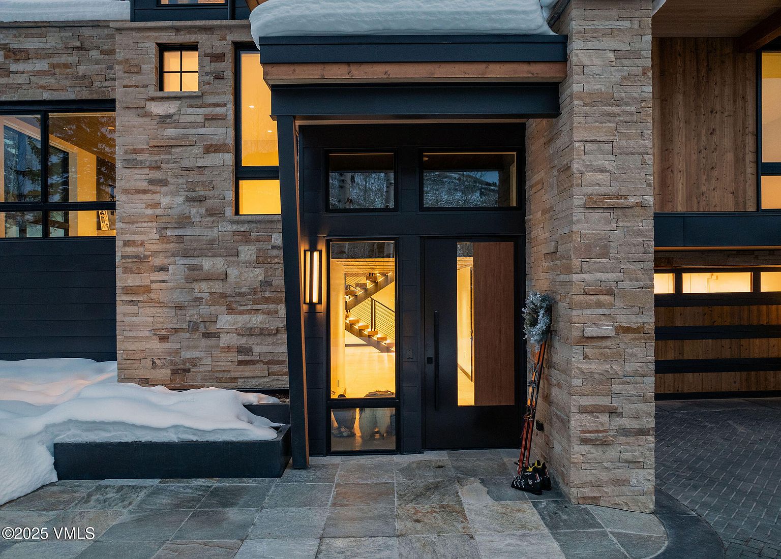 This image showcases the entryway of a modern home, featuring a stone facade and a striking black double door. The entrance is flanked by stone pillars and illuminated by contemporary light fixtures. A set of skis leans against the wall, adding a touch of mountain charm, while snow accents the landscape, creating a welcoming winter scene.