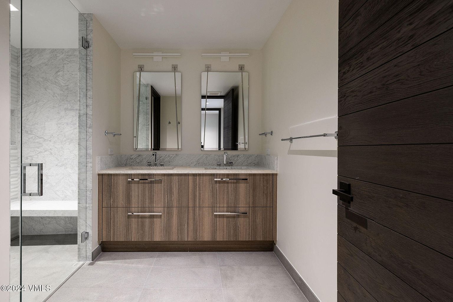 This is a well-lit primary bathroom featuring a double vanity with a light-colored countertop and wood-grain cabinets. Two mirrors hang above the sinks, each with its own light fixture. A glass-enclosed shower is visible to the left, and a dark wood door is on the right, creating a modern and clean aesthetic.
