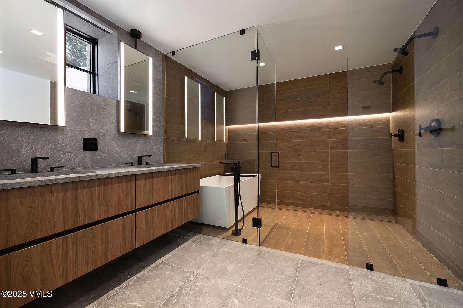This is a modern primary bathroom featuring a double vanity with a wood-grain cabinet and gray countertop, illuminated mirrors, and a glass-enclosed shower and tub area. The shower area has wood-look tile and multiple shower heads, while the floor is covered in gray tile. The perspective is a wide shot, showcasing the entire space and its luxurious features.