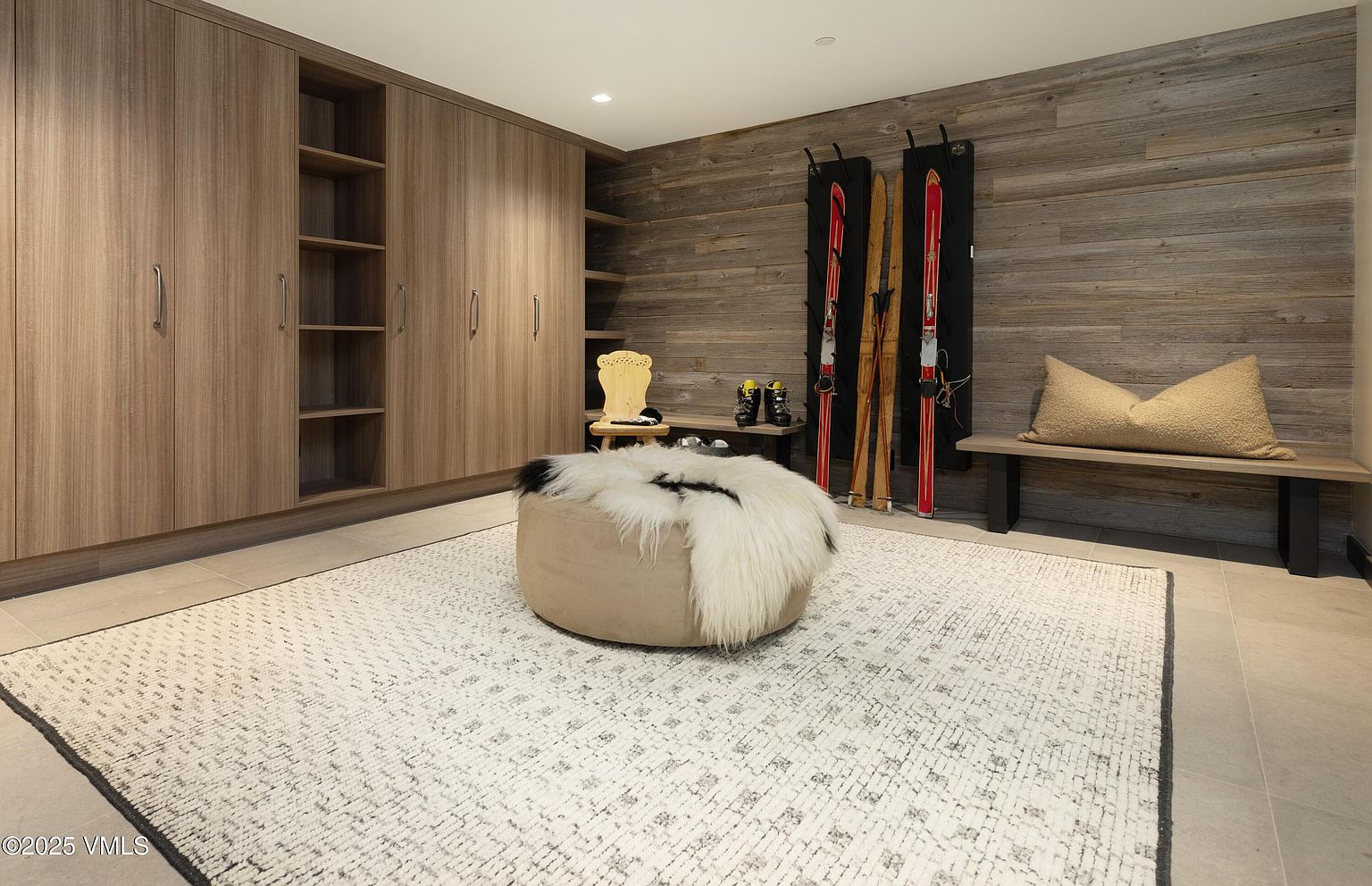 This interior shot showcases a well-organized mudroom or entryway, featuring custom wood cabinetry with ample storage. A cozy, textured rug sits in the center of the room, complemented by a round ottoman with a faux fur throw. Ski equipment is neatly arranged against the wood-paneled wall, adding a touch of rustic charm.