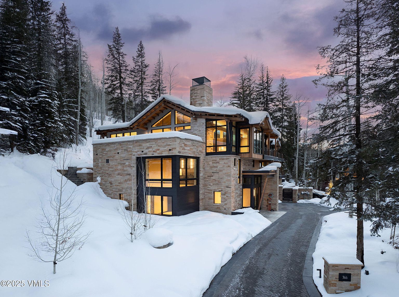 This is a front exterior view of a modern luxury home in a snowy, wooded setting. The house features a stone facade, large windows with dark frames, and a unique roofline. A paved driveway leads up to the house, and the surrounding landscape is covered in snow, with tall evergreen trees visible in the background.