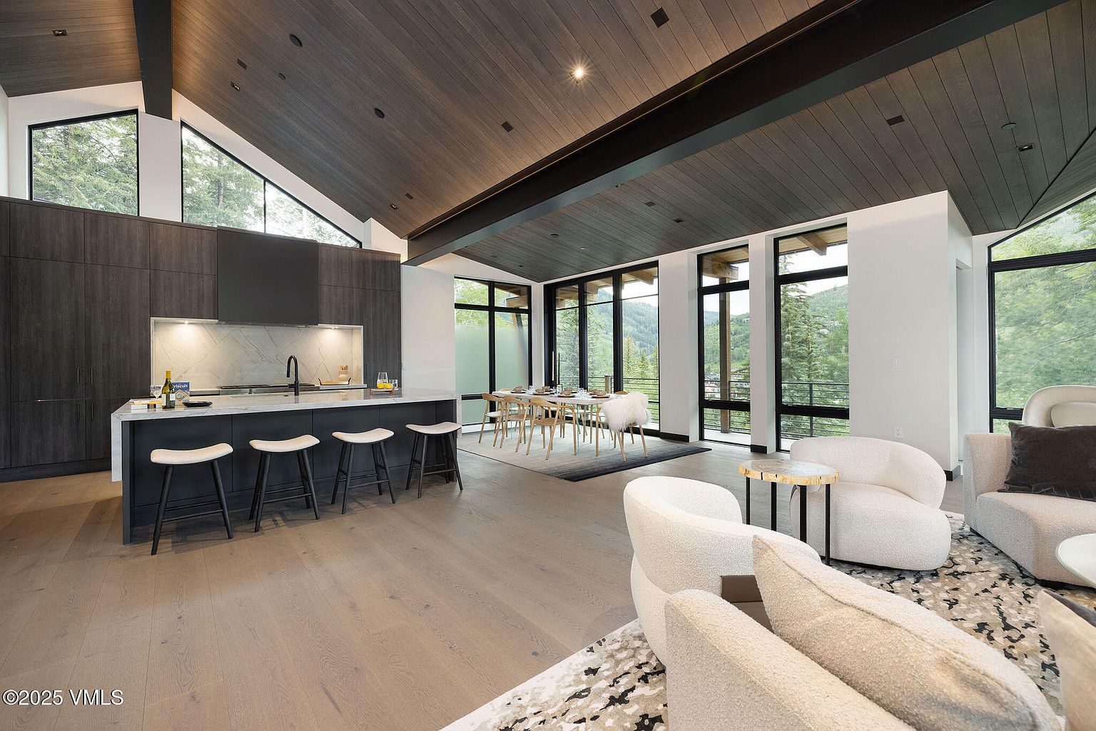 This is a spacious, modern kitchen and dining area featuring dark wood cabinetry, a large island with seating, and an open floor plan. The room is well-lit with natural light from large windows offering views of the surrounding landscape. The overall impression is one of luxury and contemporary design.