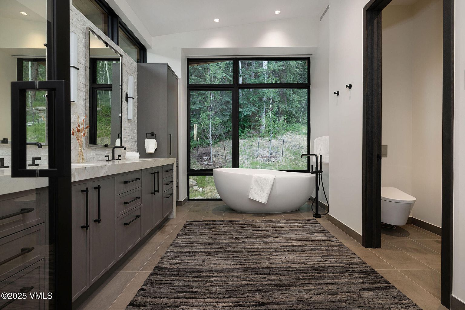 This is a modern primary bathroom featuring a large window overlooking a forest, a freestanding bathtub, and a long vanity with gray cabinets and a marble countertop. The room has a neutral color palette with black accents, creating a spa-like atmosphere. The perspective is from the entrance of the bathroom, showcasing the rug and the layout of the room.