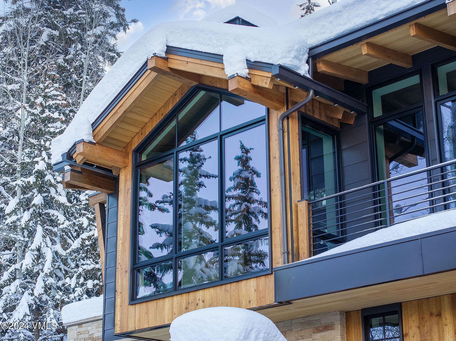 This is a side view of a modern home showcasing a large, triangular window reflecting snow-covered trees. The exterior features a combination of wood and dark gray siding, complemented by a metal balcony railing. The roof is covered in snow, adding to the wintery aesthetic.