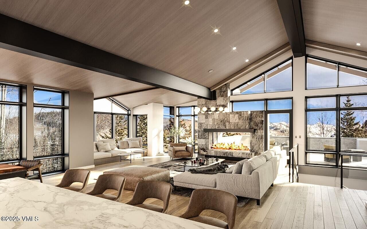 This is a spacious living room featuring a stone fireplace, large windows with mountain views, and modern furnishings. The room has a high, vaulted ceiling with wooden beams, creating an open and airy feel. The neutral color palette and natural light contribute to a sophisticated and inviting atmosphere.