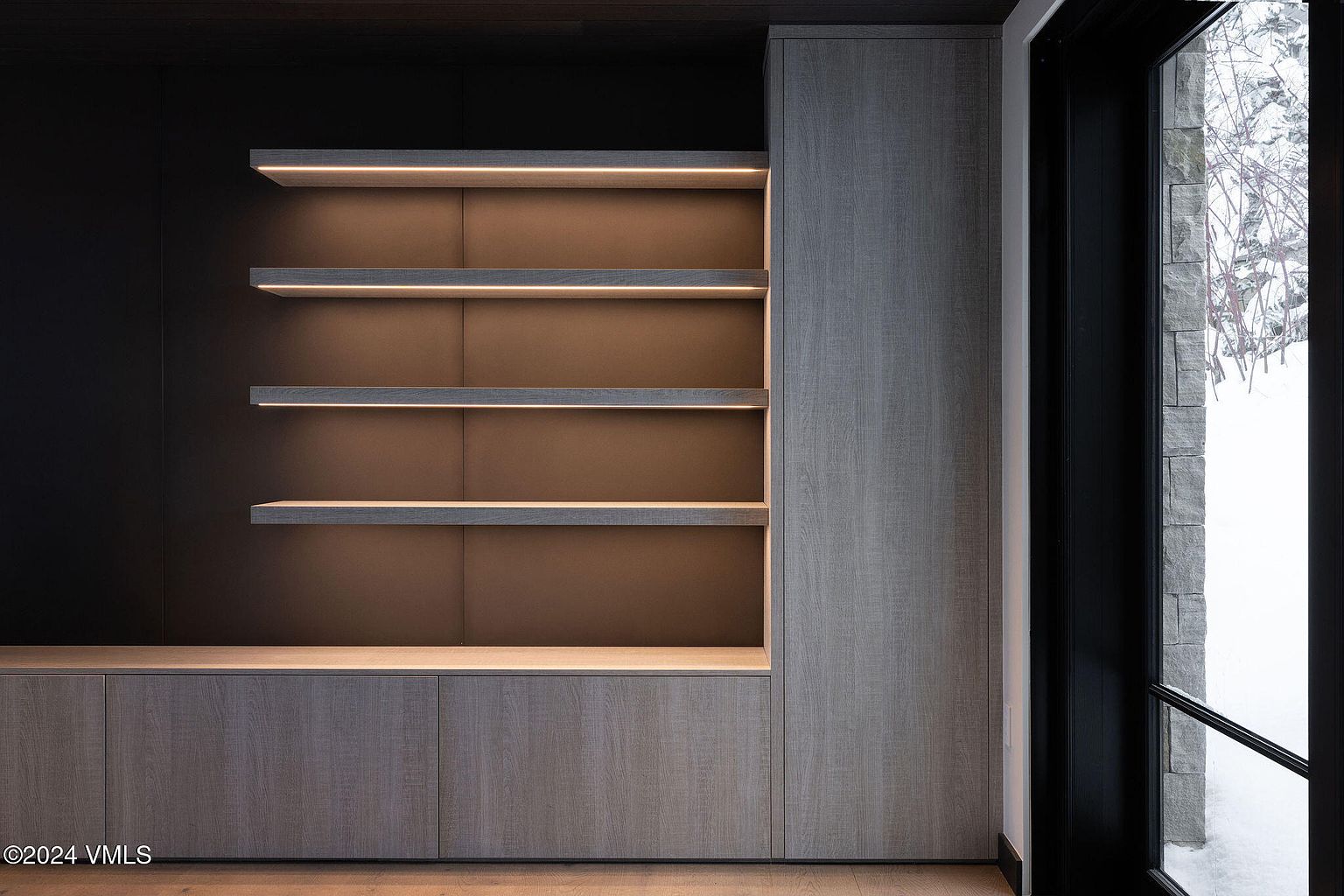 This image showcases a modern living room with a focus on built-in shelving and cabinetry. The shelves are illuminated with subtle lighting, creating a warm ambiance. A large window provides natural light and a view of the outdoors, enhancing the room's appeal.