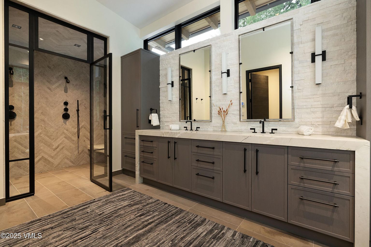 This is a primary bathroom featuring a double vanity with gray cabinetry and black hardware, topped with a light-colored countertop. Two mirrors hang above the sinks, flanked by modern sconces, and a glass-enclosed shower is visible to the left. The overall design is contemporary and luxurious, with a focus on clean lines and neutral tones.
