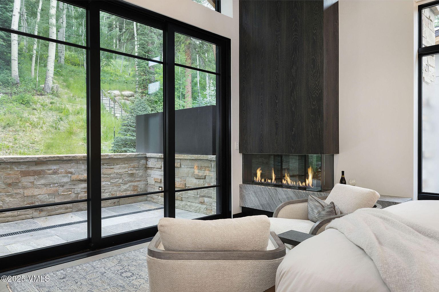 This is a primary bedroom featuring a modern fireplace with a dark wood surround and glass enclosure. Large windows offer a view of the outdoors, and the room is furnished with comfortable seating and a bed. The overall style is contemporary and luxurious.