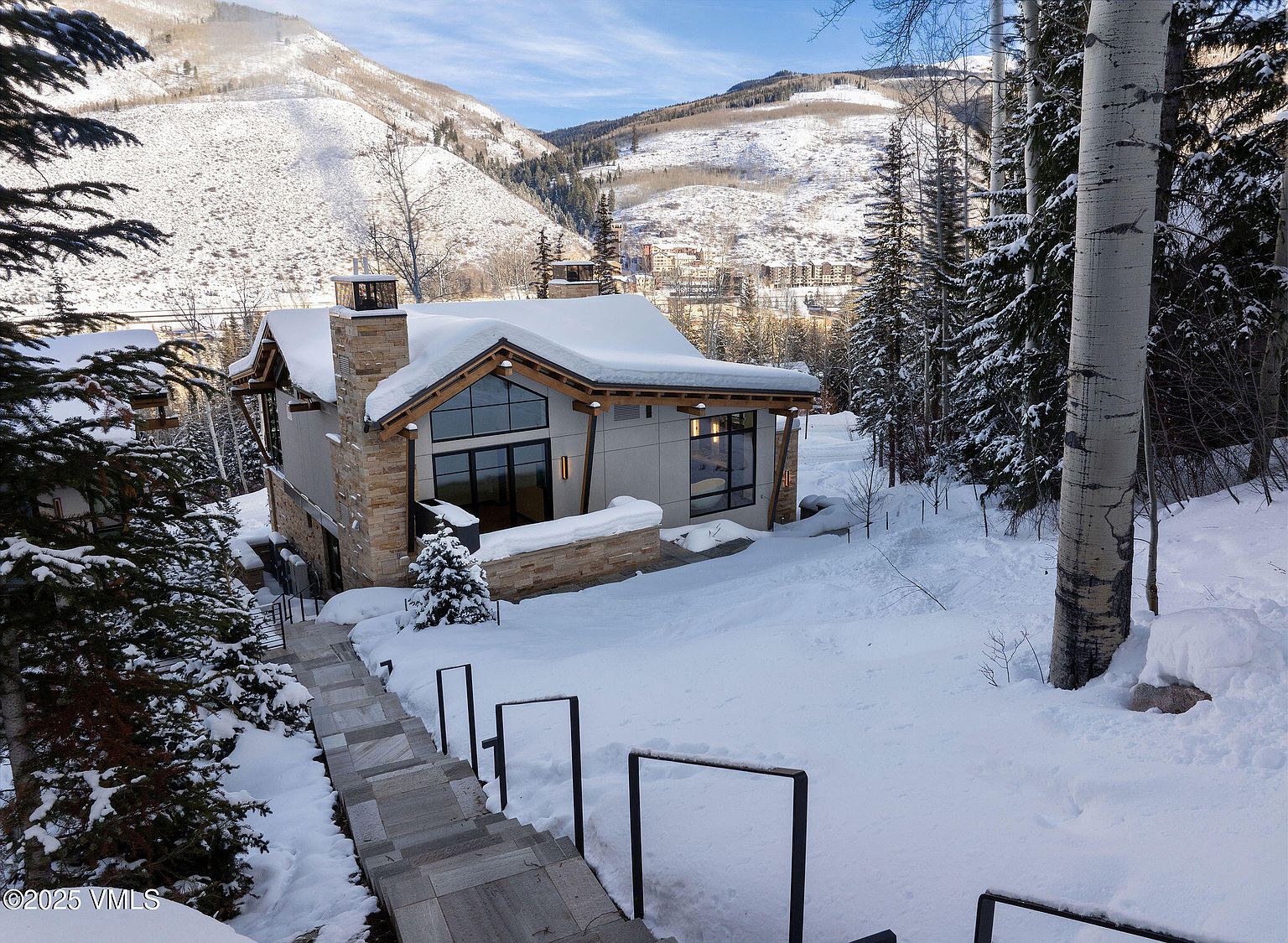 This image showcases the front exterior of a modern mountain home in a snowy setting. The house features a combination of stone and light-colored siding, large windows, and a snow-covered roof. A set of stairs leads up to the house, and the surrounding landscape includes snow-covered trees and mountains in the background, creating a serene and luxurious winter retreat.