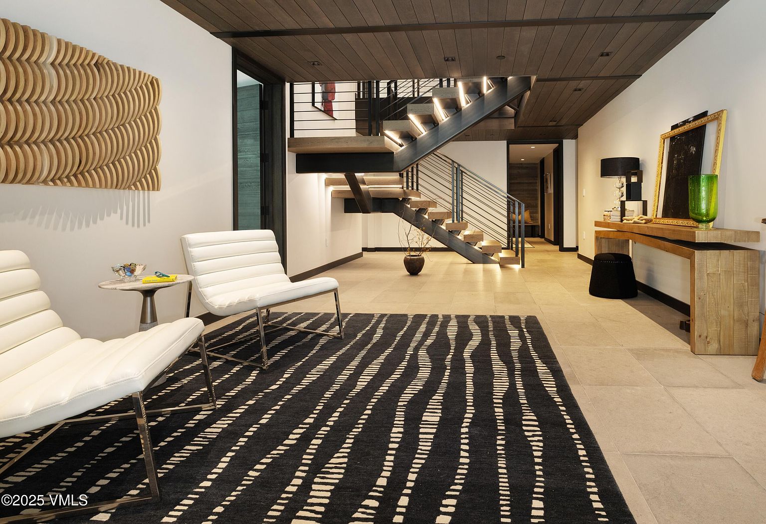 This interior shot showcases a modern hallway and staircase. The staircase features open wooden treads with metal railings and integrated lighting. The hallway includes contemporary furnishings such as white chairs, a black and white patterned rug, and a wooden console table, creating a stylish and inviting entrance area.
