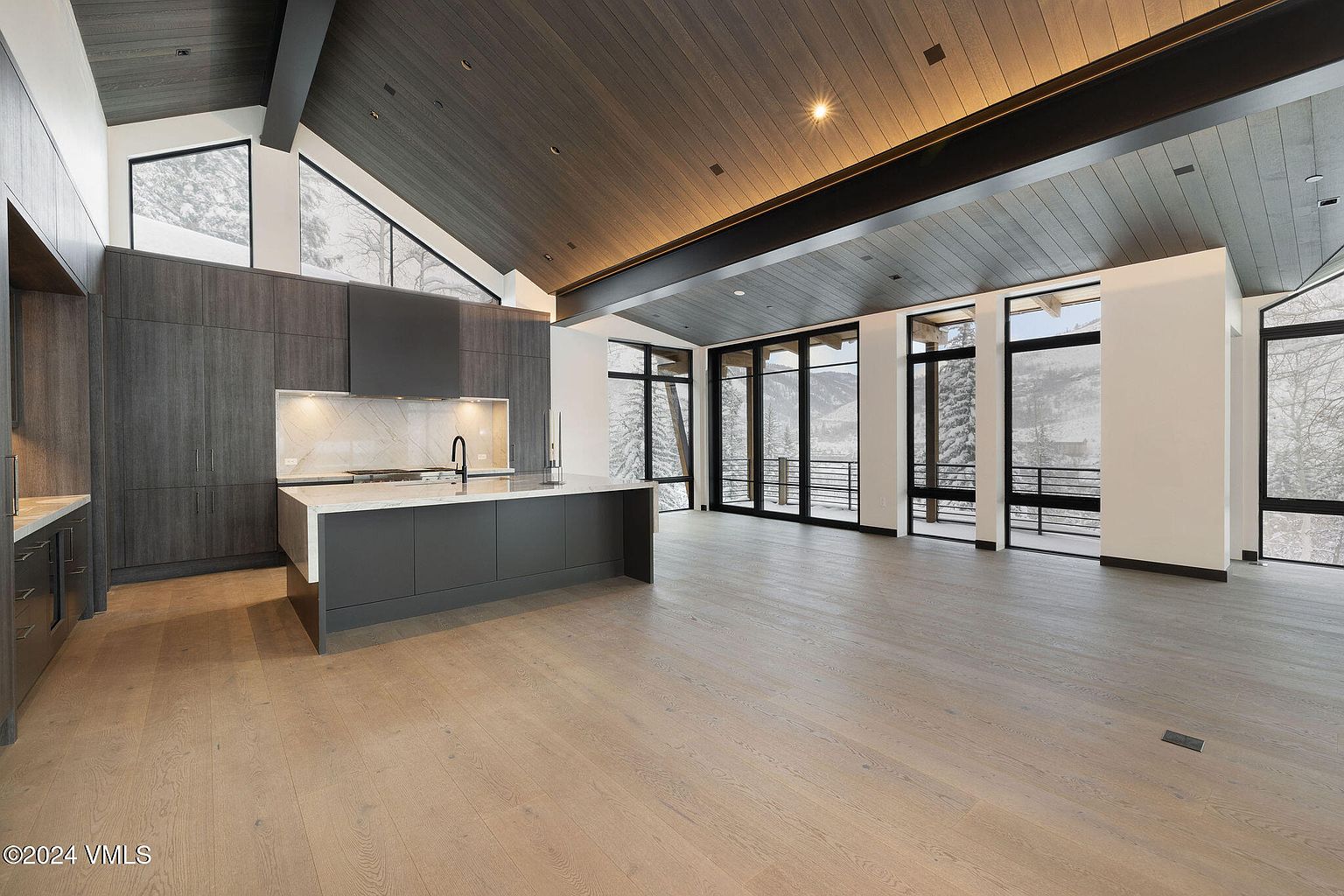 This interior shot showcases a modern kitchen and open living space with a focus on natural light and clean lines. The kitchen features dark wood cabinetry, a large island with a white countertop, and stainless steel appliances. Expansive windows offer views of a snowy landscape, enhancing the contemporary and luxurious feel of the home.