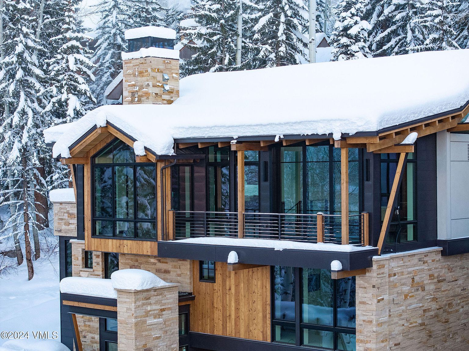 This is a front exterior view of a modern mountain home covered in snow. The house features a combination of stone and wood siding, large windows, and a balcony with metal railings. The surrounding landscape is filled with snow-covered evergreen trees, creating a serene and luxurious winter setting.