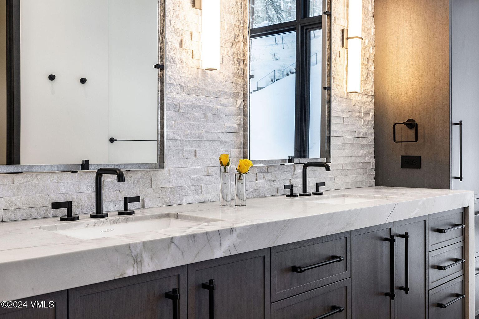 This is a well-lit primary bathroom featuring a double vanity with a marble countertop and dark gray cabinetry. The backsplash is made of stacked stone, and the mirrors are framed in black. Modern black fixtures and hardware complement the contemporary design, creating a luxurious and stylish space.