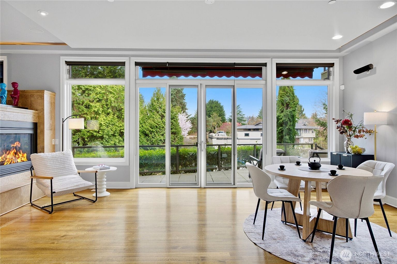 This bright and airy living space features light wood flooring and a cozy seating area arranged around a fireplace with a tall stone-clad surround. A large dining set sits on a circular rug to the right, while expansive glass sliding doors provide a seamless transition to the outdoor deck and offer panoramic views of the surrounding greenery. The room is illuminated by recessed lighting and a floor lamp, creating a warm and inviting atmosphere.