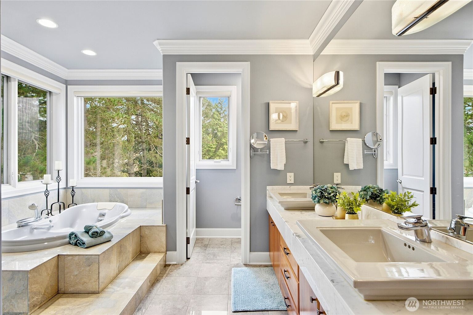 This primary bathroom features a large soaking tub set into a tiled platform beside a window with a view of the trees. A double vanity with a stone countertop and dual sinks sits opposite the tub, complemented by a large wall-to-wall mirror and modern light fixtures. The space is finished in a neutral color palette with light-toned tile flooring and soft gray walls, creating a bright and serene atmosphere.