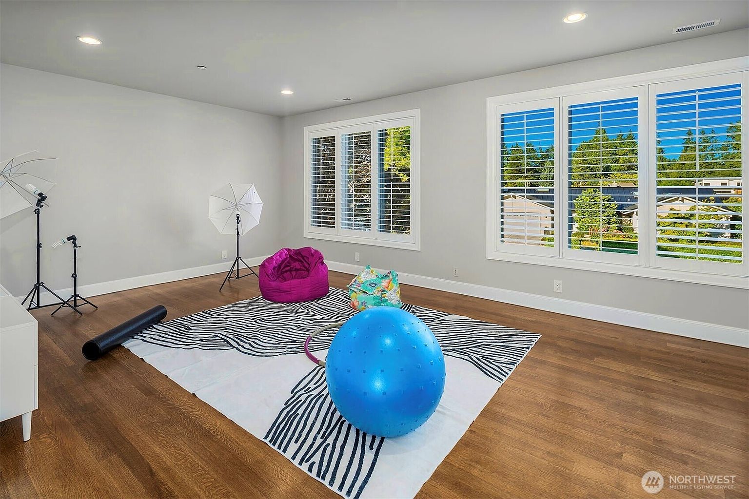 This bright, multi-purpose room features warm-toned wood flooring and light-colored walls, creating an inviting atmosphere. The space is currently set up as a home gym or studio, equipped with a large blue exercise ball, a yoga mat, and photography lighting equipment. Large windows with white shutters allow for ample natural light, providing a pleasant view of the neighborhood outside.