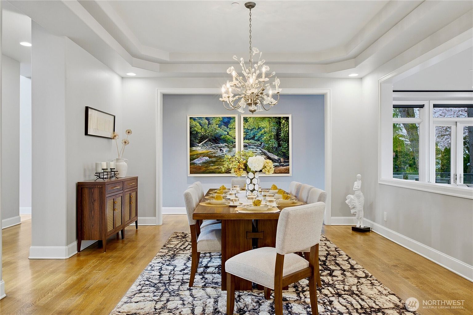 This elegant dining room features a central wooden table surrounded by six upholstered chairs, set atop a large, patterned area rug. A sophisticated crystal chandelier hangs from a tray ceiling, while a wooden sideboard with cane-front doors sits against the left wall. The space is accented by two large landscape paintings on a soft blue feature wall and a white sculptural figure, creating a refined and welcoming atmosphere.