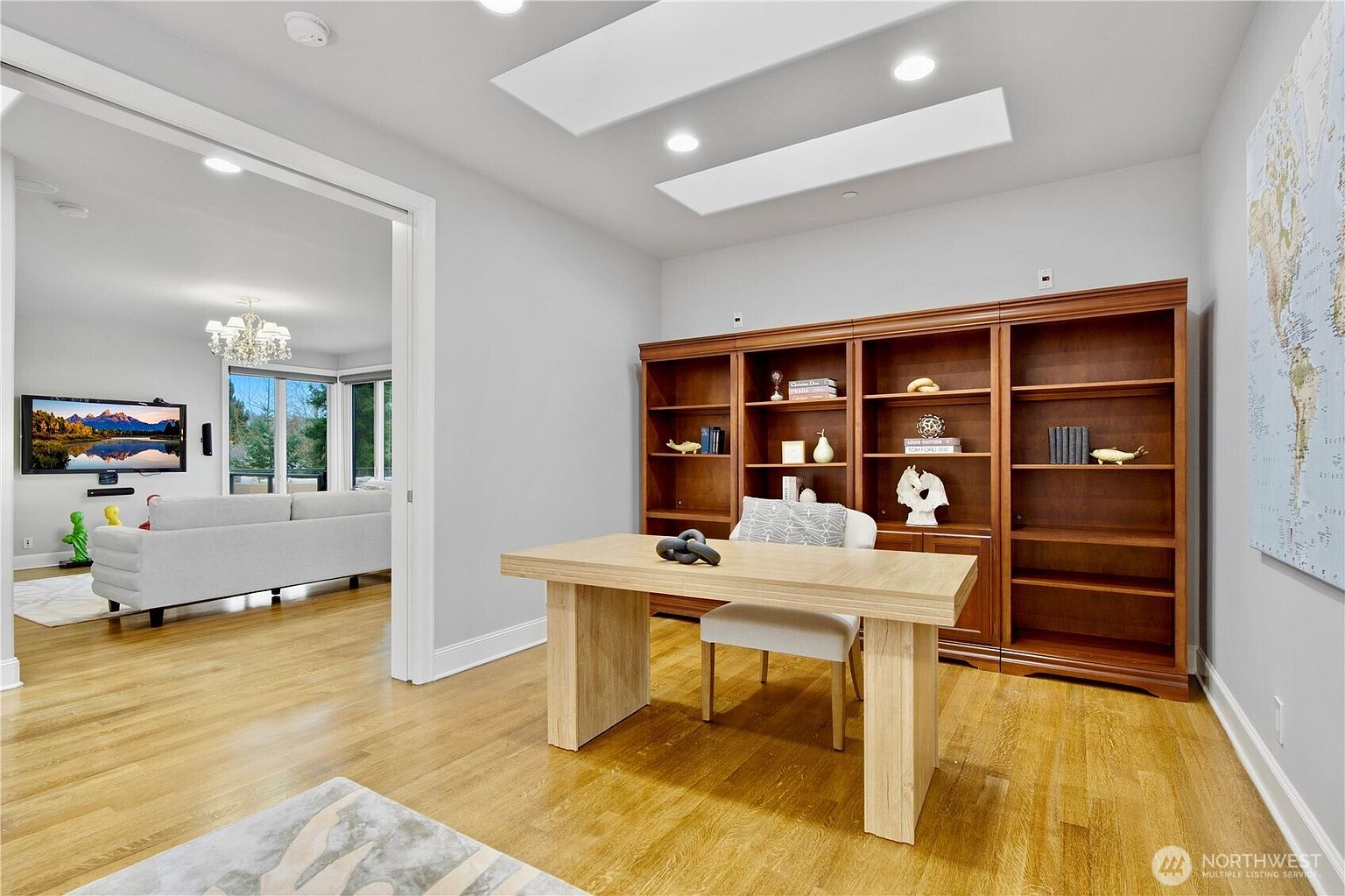 This bright home office features light wood flooring and a minimalist desk centered in front of a large, built-in wooden bookshelf. The room is illuminated by recessed lighting and two rectangular skylights, creating an airy and professional atmosphere. A wide doorway provides a clear view into an adjacent living area, showcasing a cohesive and open floor plan.