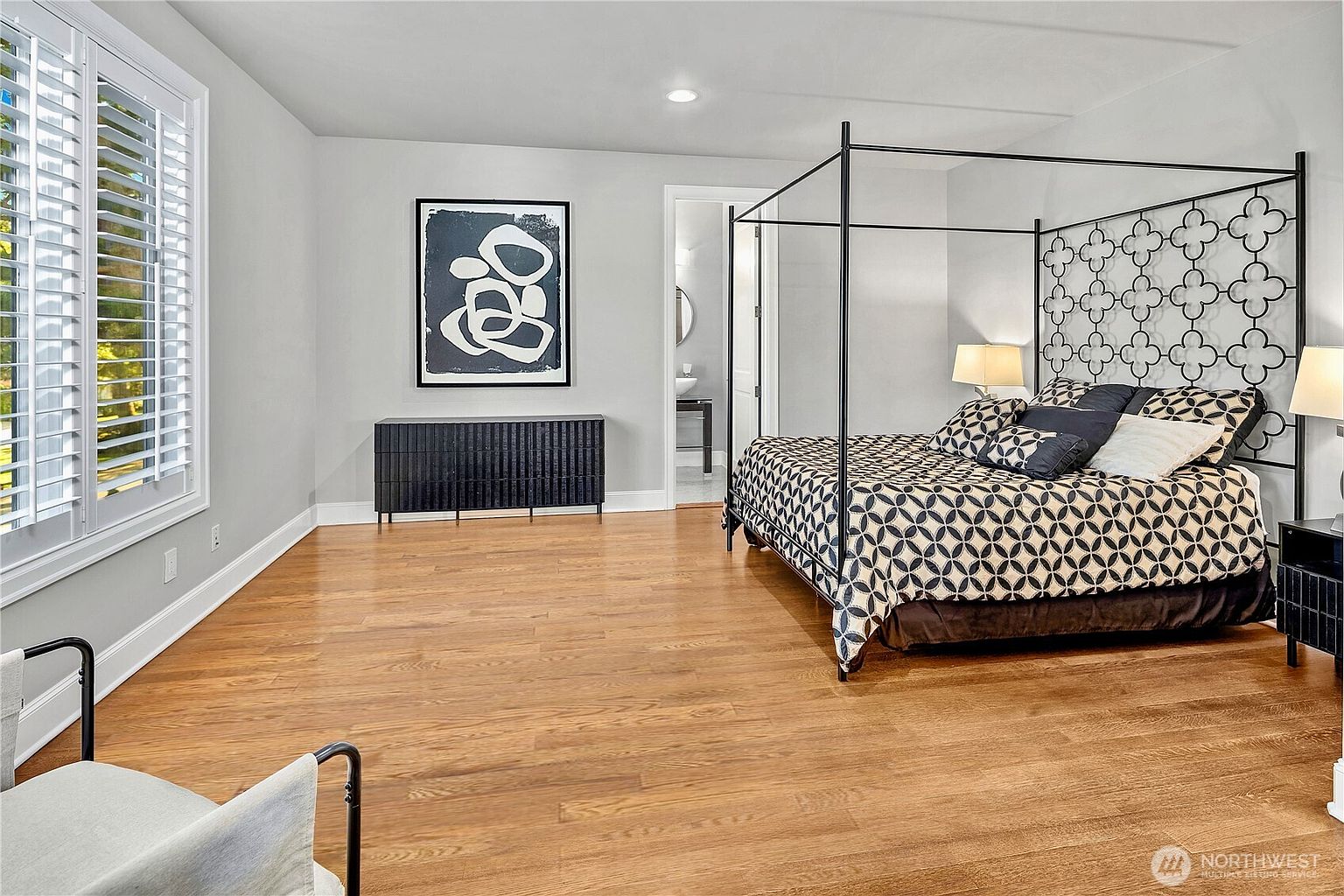 This spacious primary bedroom features warm-toned wood flooring and light-colored walls, creating a bright and airy atmosphere. A prominent black metal canopy bed with a geometric patterned headboard serves as the focal point, complemented by a matching black dresser and abstract wall art. Large windows with white shutters allow natural light to fill the room, which also includes a doorway leading to an adjoining bathroom.